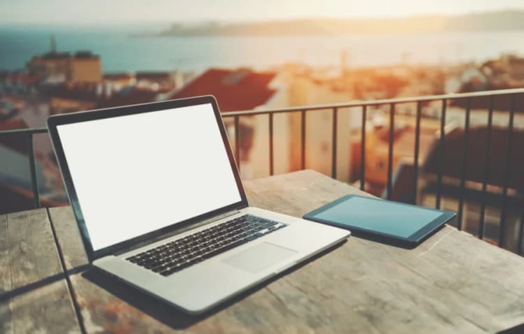 A laptop and tablet lying on a desk on a balcony with a view of Lisbon behind them